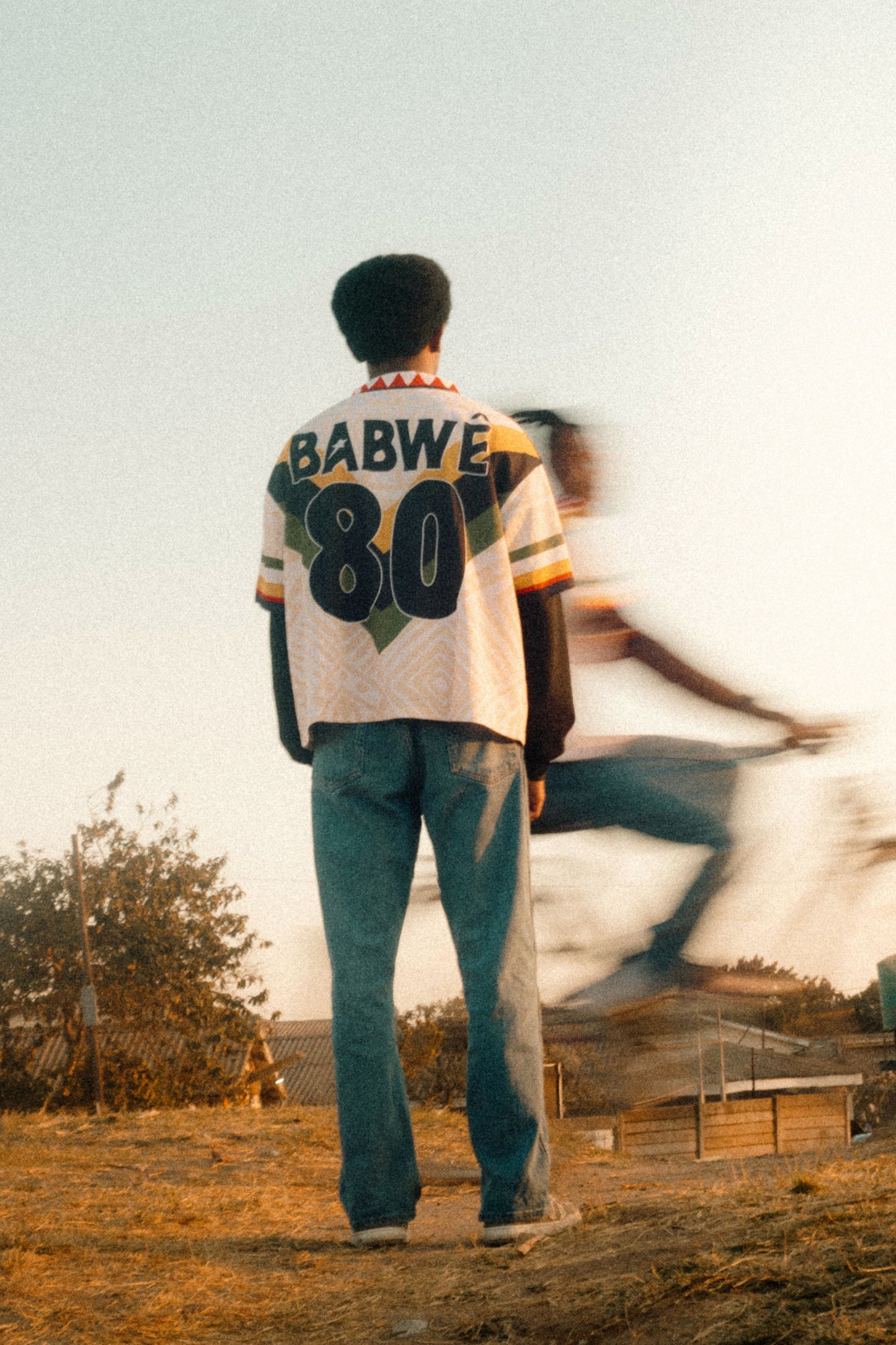 Person wearing a Vintage Zimbabwean soccer jersey featuring national colors and retro design – inspired by classic 90s football culture standing in an open field with another person running in the background.