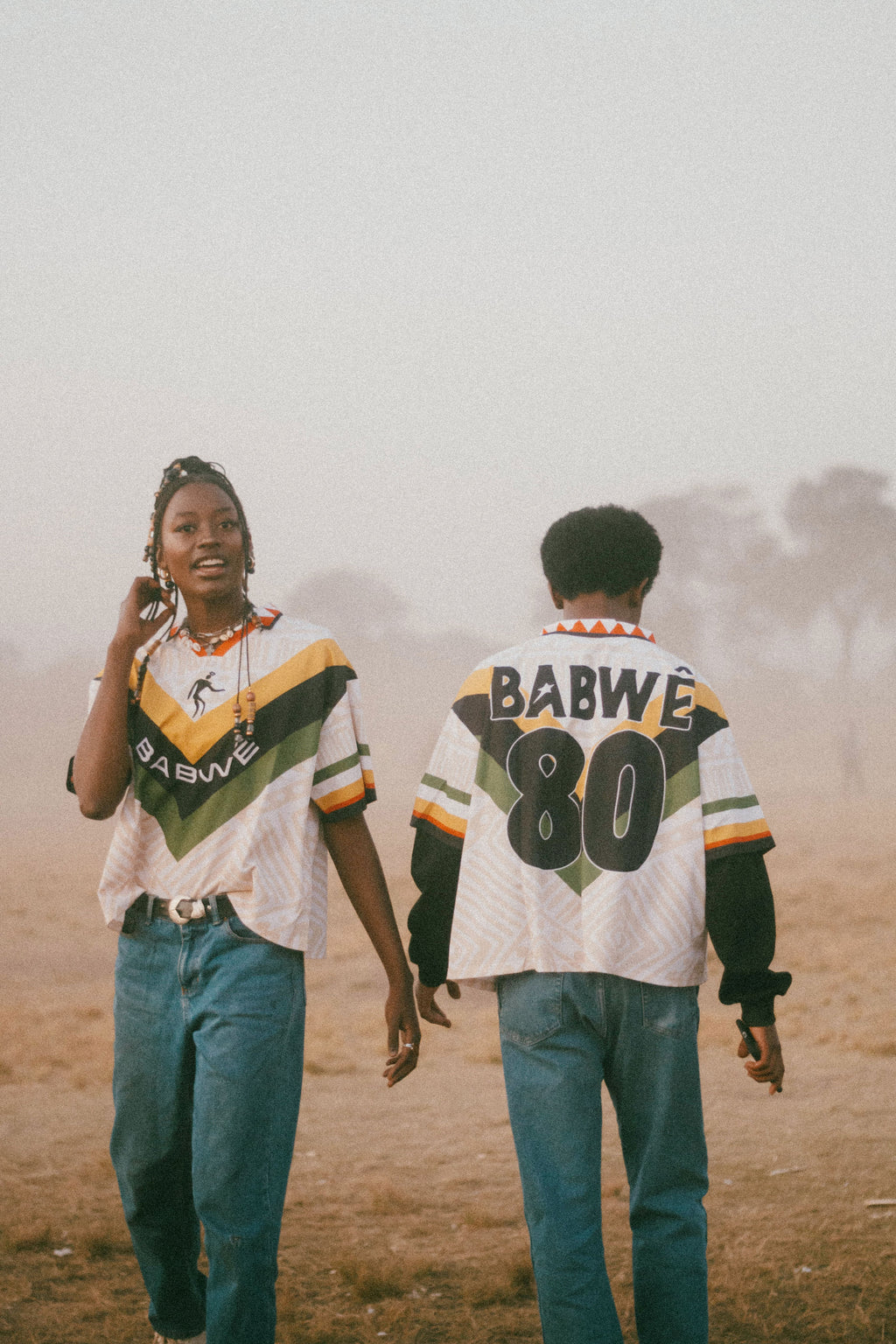 Two people wearing matching Babwê vintage Zimbabwe soccer jersey – nostalgic 90s design blending African streetwear with national pride on a misty field.